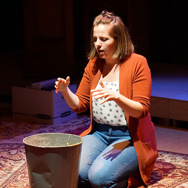 woman in jeans and red sweater kneels on floor, gesturing over a trash can