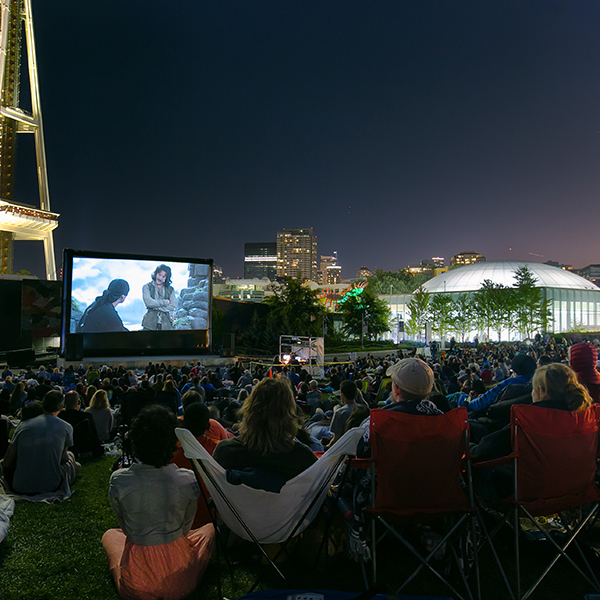 a movie screen glows in darkness while patrons sitting in lawn chairs watch