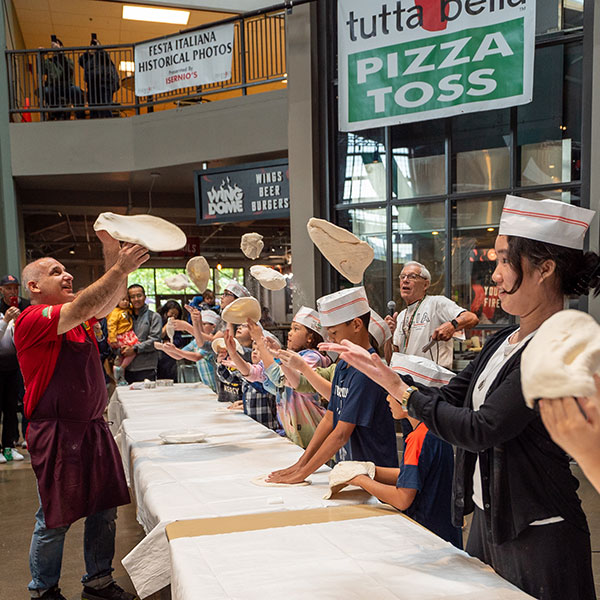 Man instructing a table full of children tossing pizza dough in the air.